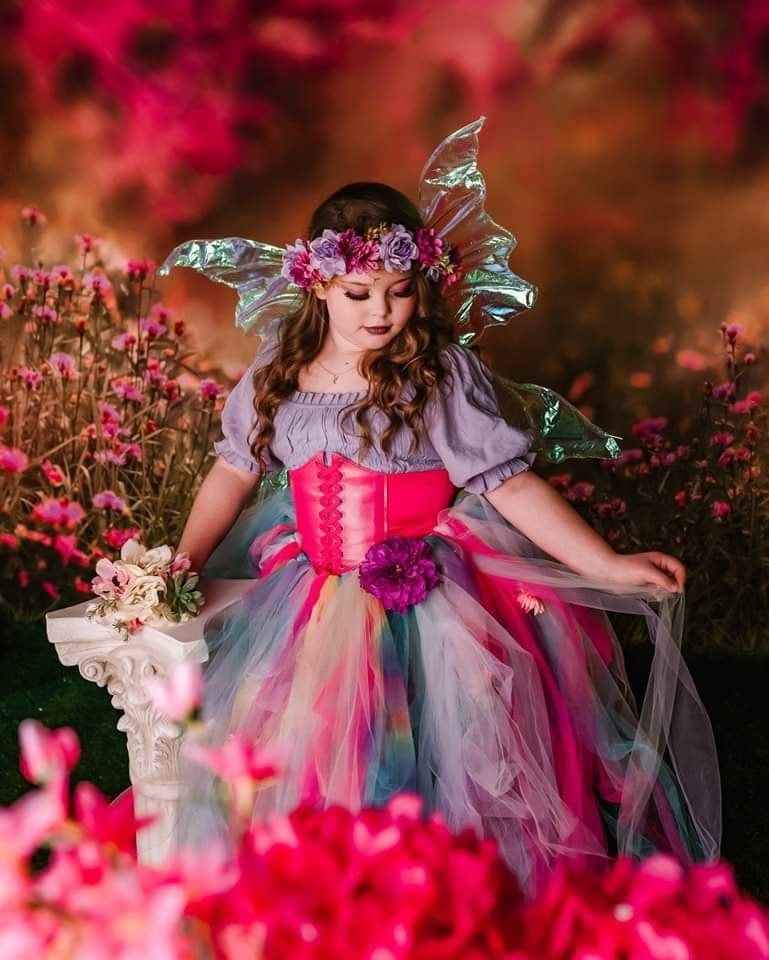 A young girl in a colorful fairy costume with tutu dress, floral crown, and wings poses among pink flowers.