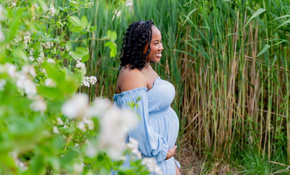 Pregnant woman in light blue off-shoulder dress standing among green reeds and white flowers outdoors.
