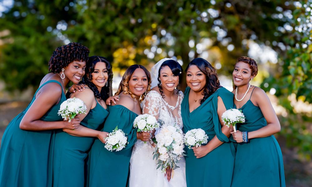 Bride in white lace gown poses with five bridesmaids in teal dresses holding white bouquets outdoors.