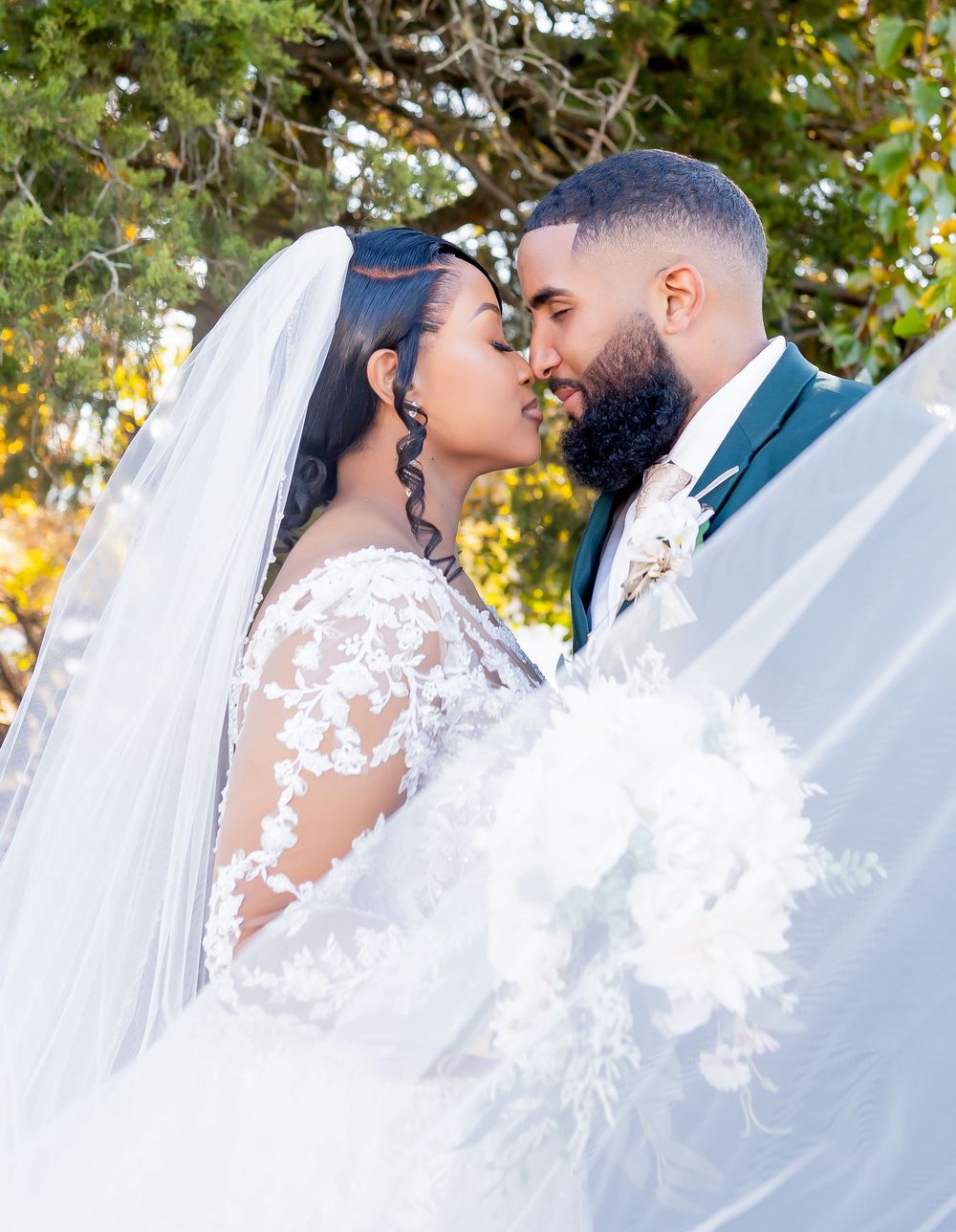 A bride and groom share a romantic nose-to-nose moment, draped in a flowing white veil, surrounded by golden autumn trees.