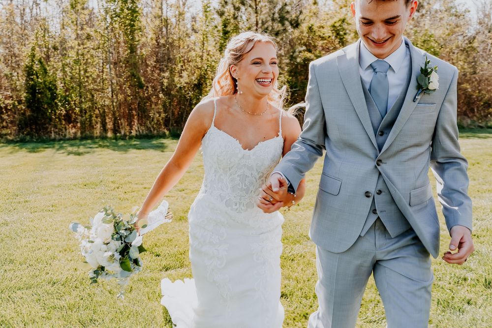 Happy bride and groom holding hands walking outdoors, bride in white gown with bouquet, groom in light gray suit.
