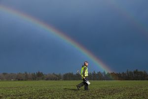 Worker in hi-vis vest walks across green field beneath a vibrant rainbow against dramatic stormy sky.