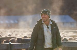 Smiling farmer in beanie and jacket stands by livestock pens on a sunny rural farm day.