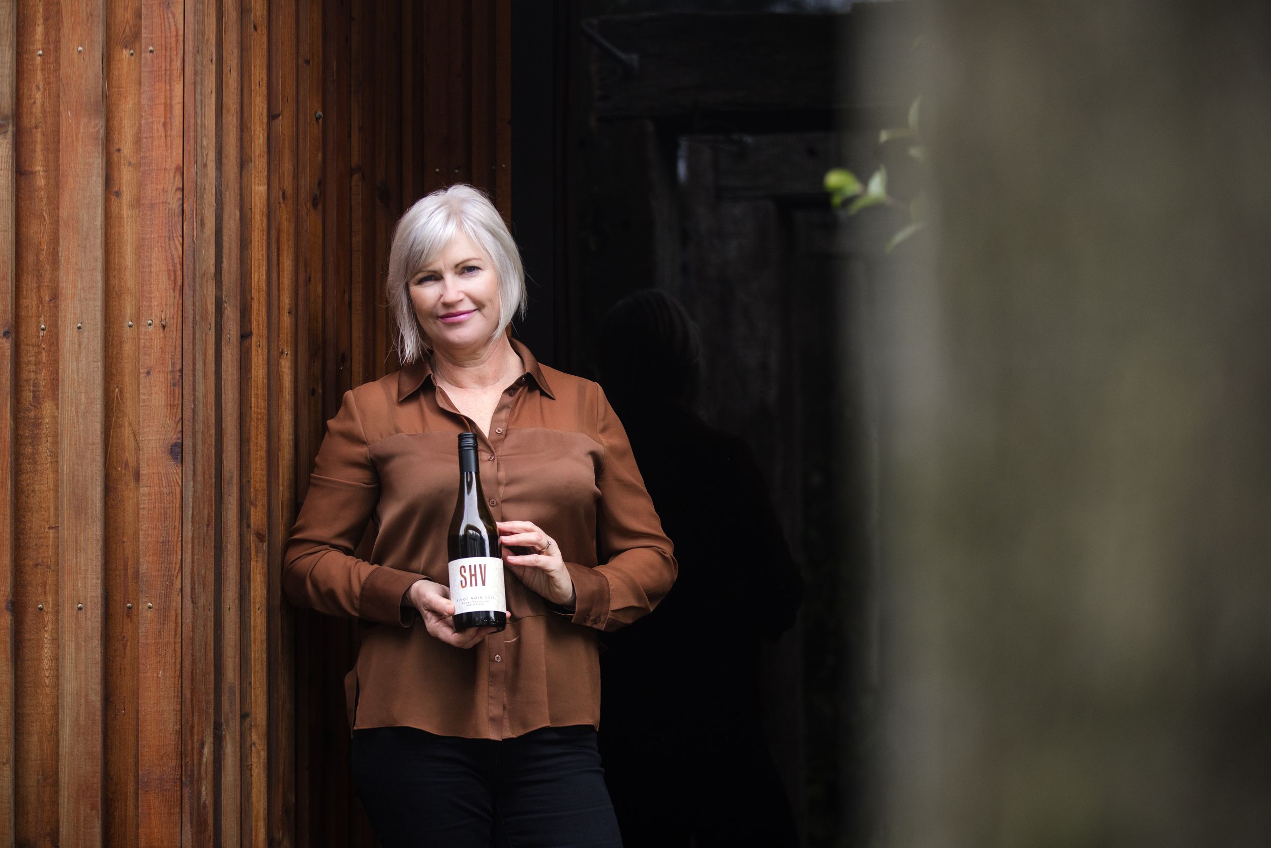 Person in brown jacket stands against rustic wooden wall while holding a wine bottle