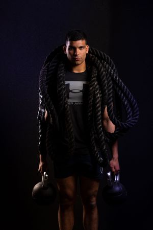Athletic figure in dark workout attire performs battle rope exercises against dramatic black background.