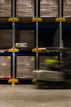 Forklift moving through a large warehouse with tall yellow shelving units stocked with cardboard boxes on pallets.