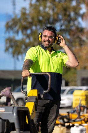 Worker in hi-vis shirt wearing ear muffs talks on radio while operating yellow pallet jack outdoors.