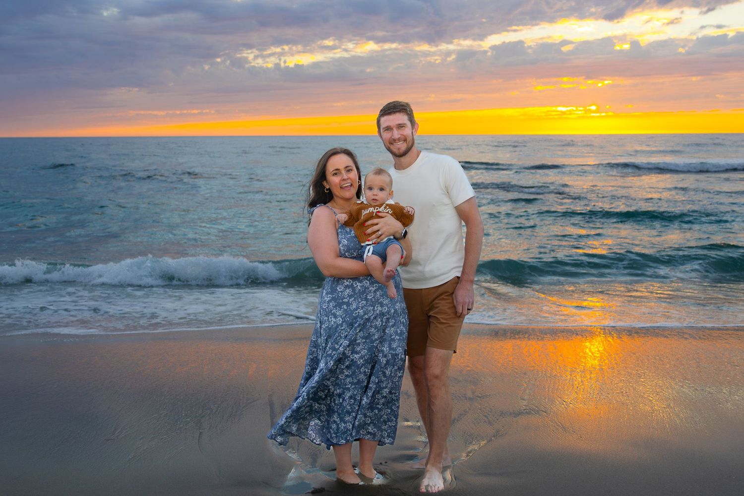 San Diego family portrait photography of a couple holding their baby on a beach at sunset with ocean waves.