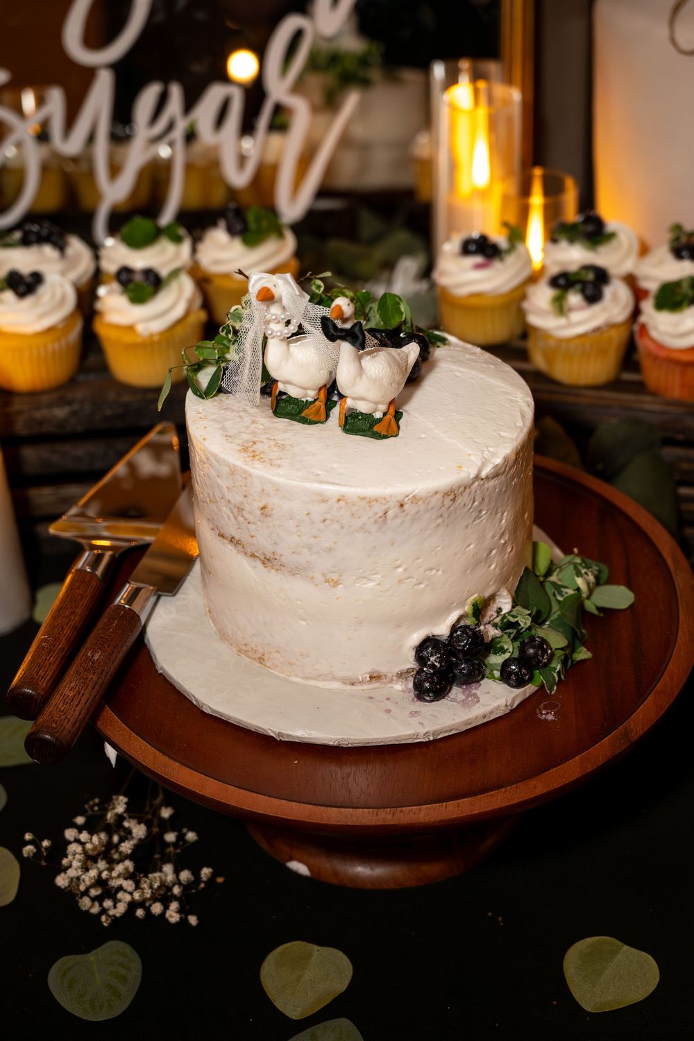 Rustic naked wedding cake with duck toppers surrounded by blueberries and greenery.