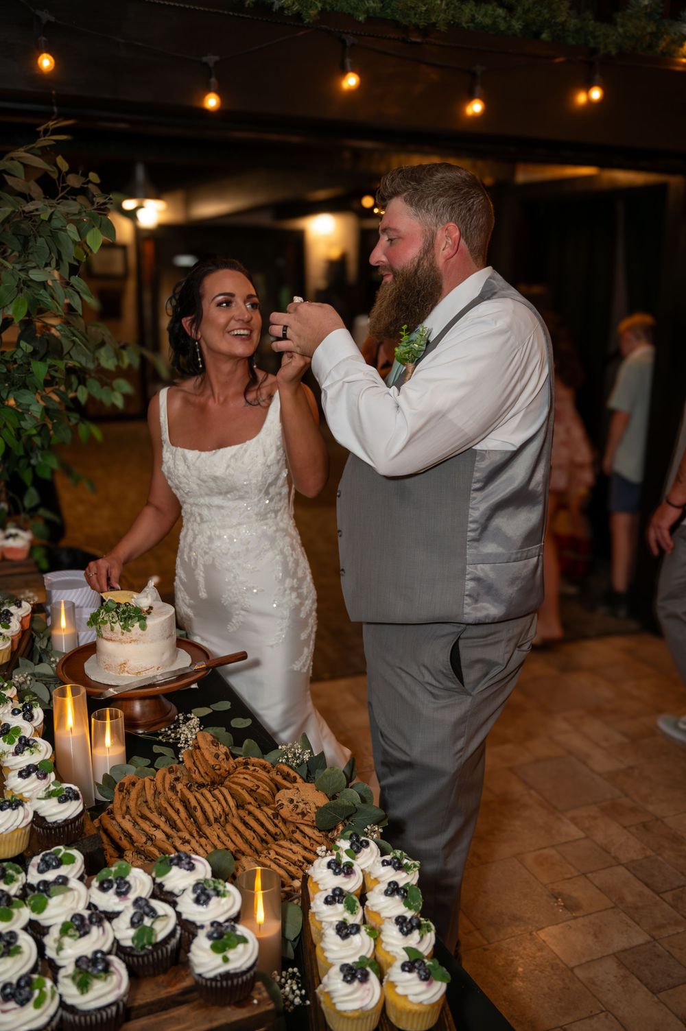 Groom feeds bride cake while smiling at dessert table with cupcakes at reception.