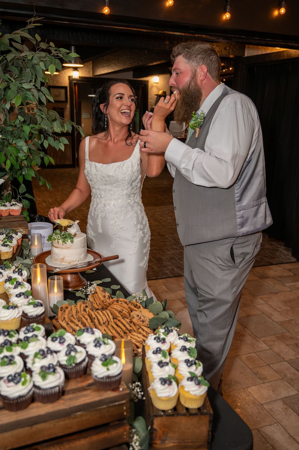 Bride feeding groom wedding cake at rustic reception with cupcake display and candles.