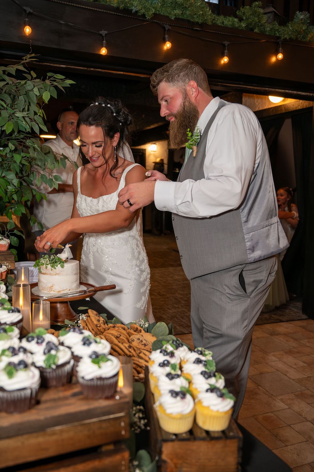 Newlyweds cutting wedding cake together at elegant reception dessert table with cupcakes.