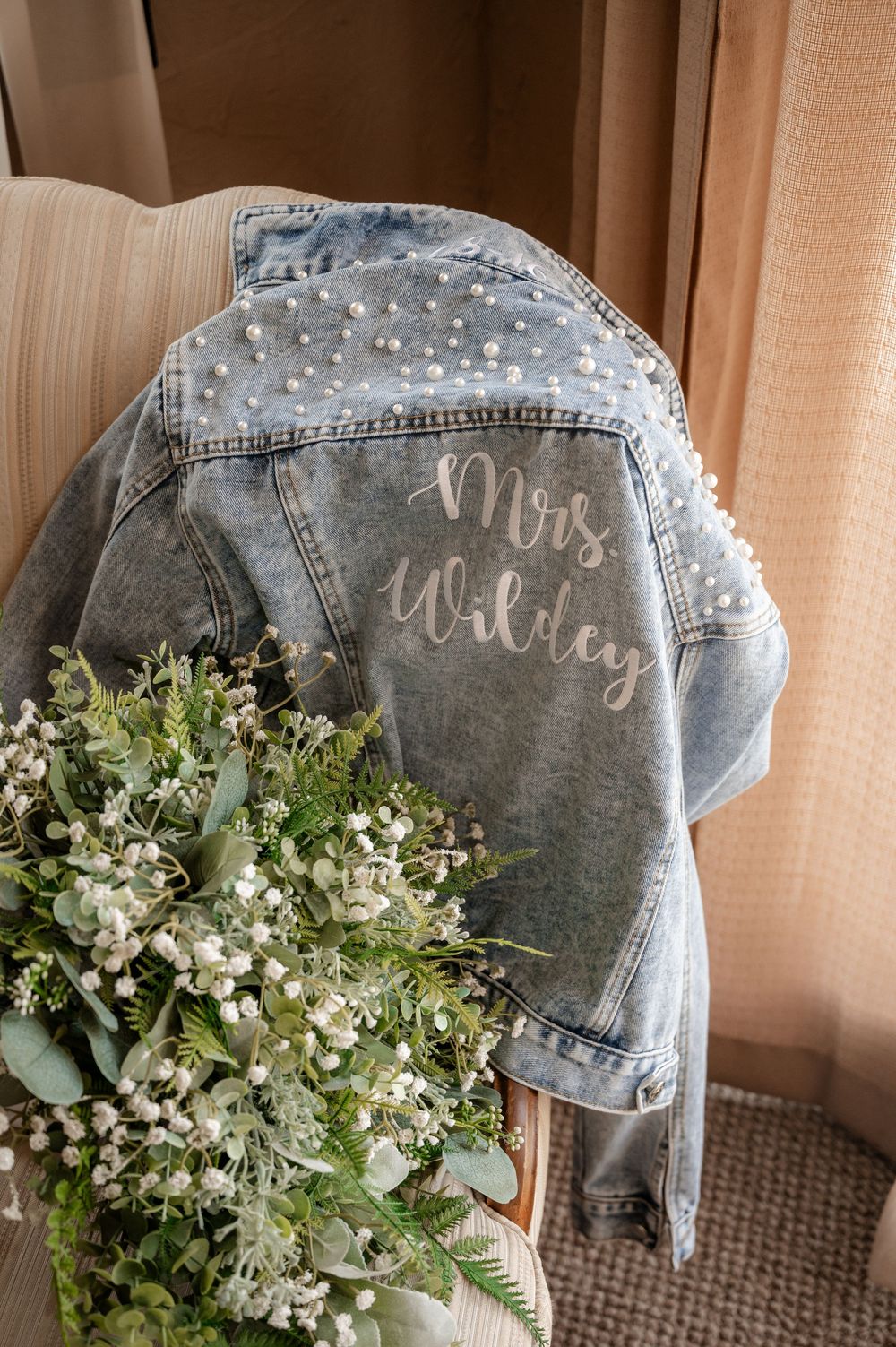 Pearl-embellished denim jacket with Mrs. Willey script beside white greenery bridal bouquet.