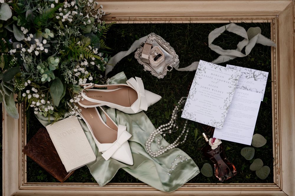 Bridal flat lay with white heels, pearl necklace, greenery bouquet, and wedding stationery.