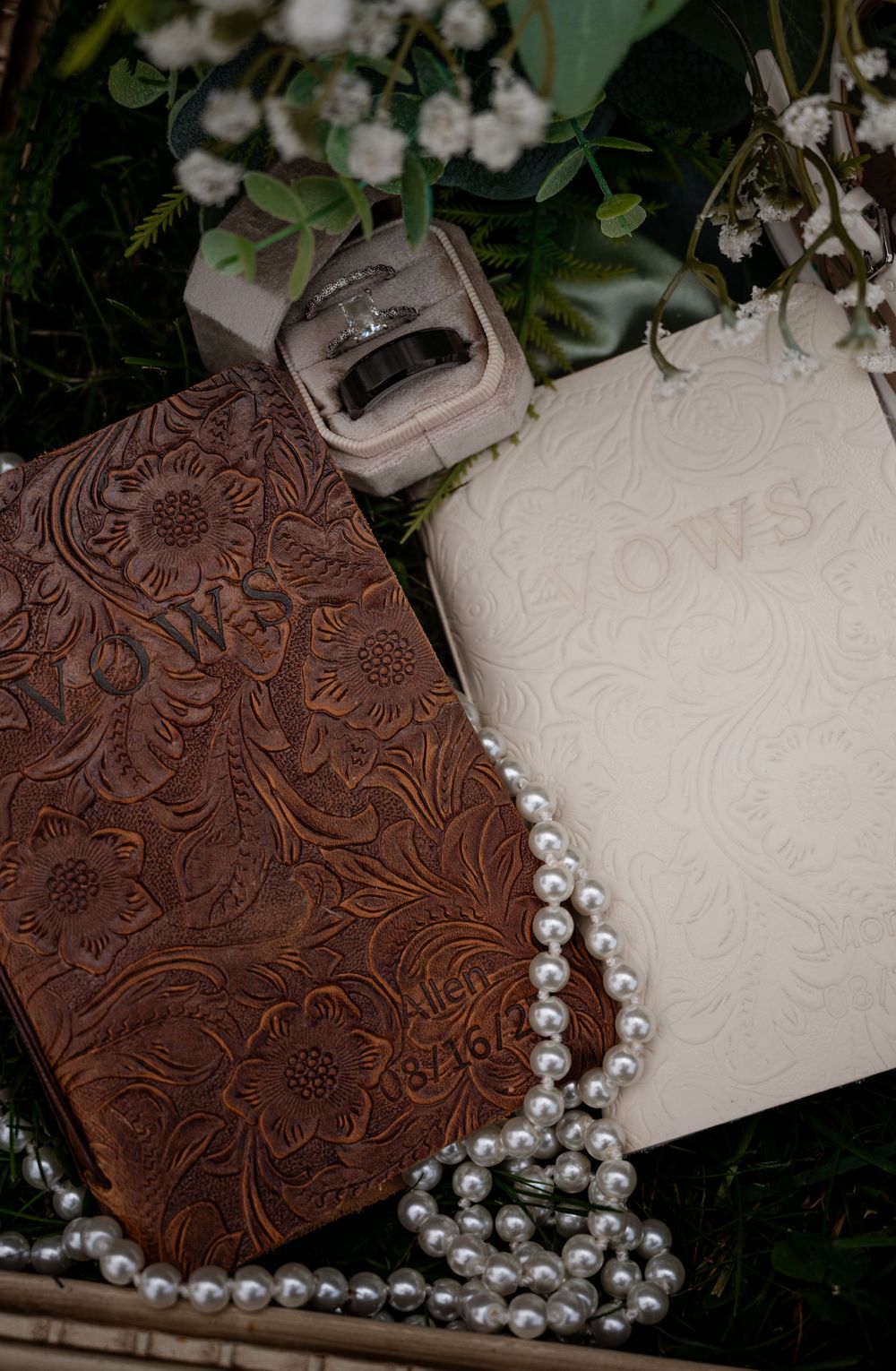 Wedding detail flat lay with tooled leather vow books, rings, and pearl necklace on greenery.