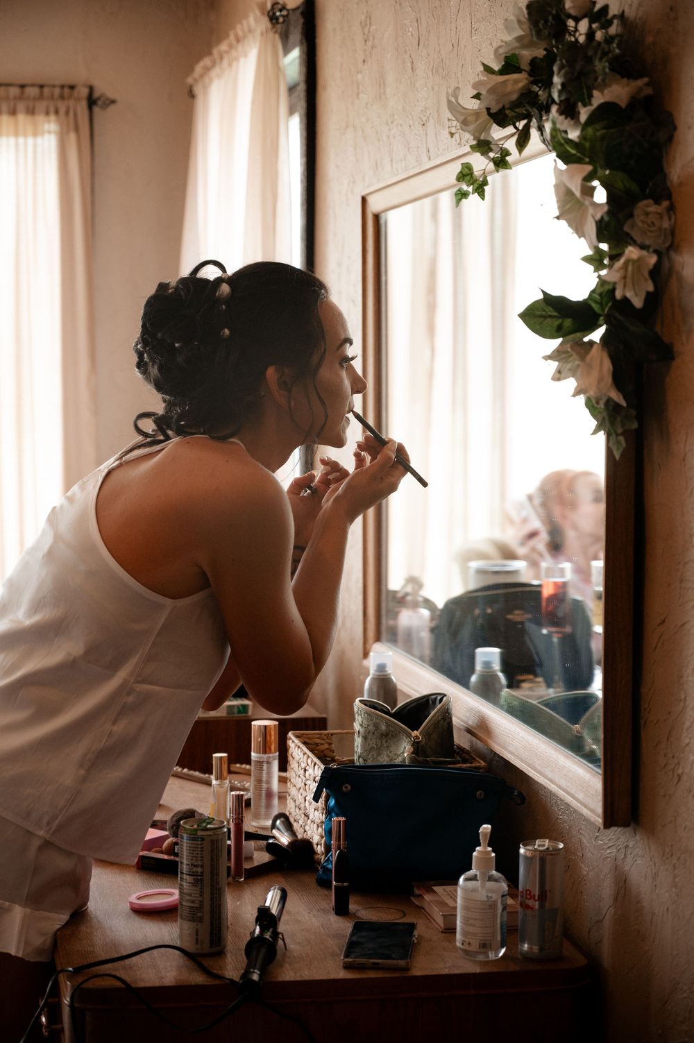 Bride applying makeup at floral-decorated mirror during wedding morning preparations.