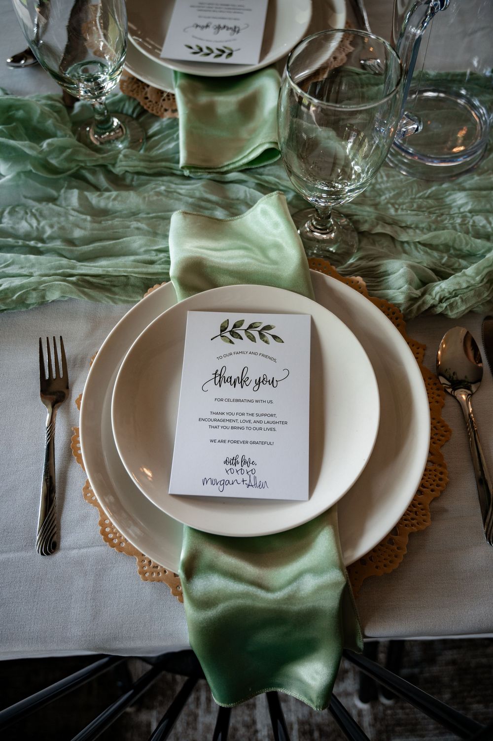 Elegant wedding place setting with sage green satin napkin and thank you card on white plates.