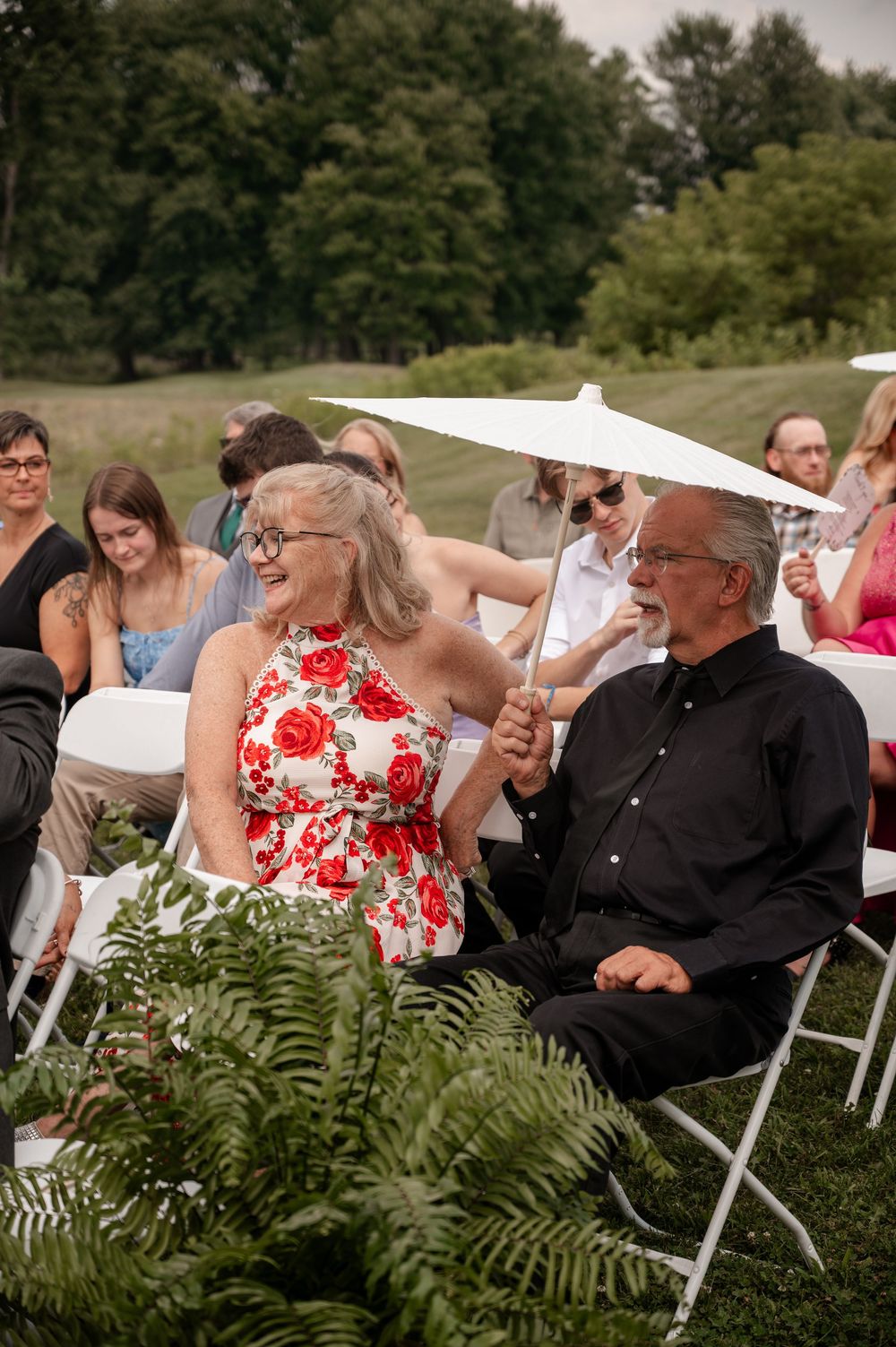Wedding guests seated outdoors on white chairs, woman in floral dress and man in black shirt smiling during ceremony.