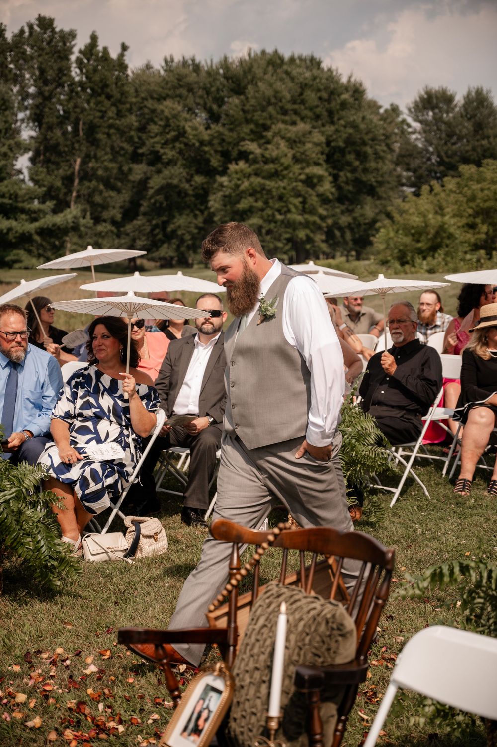 Bearded groom in gray vest and pants walking down outdoor wedding ceremony aisle with guests seated behind.