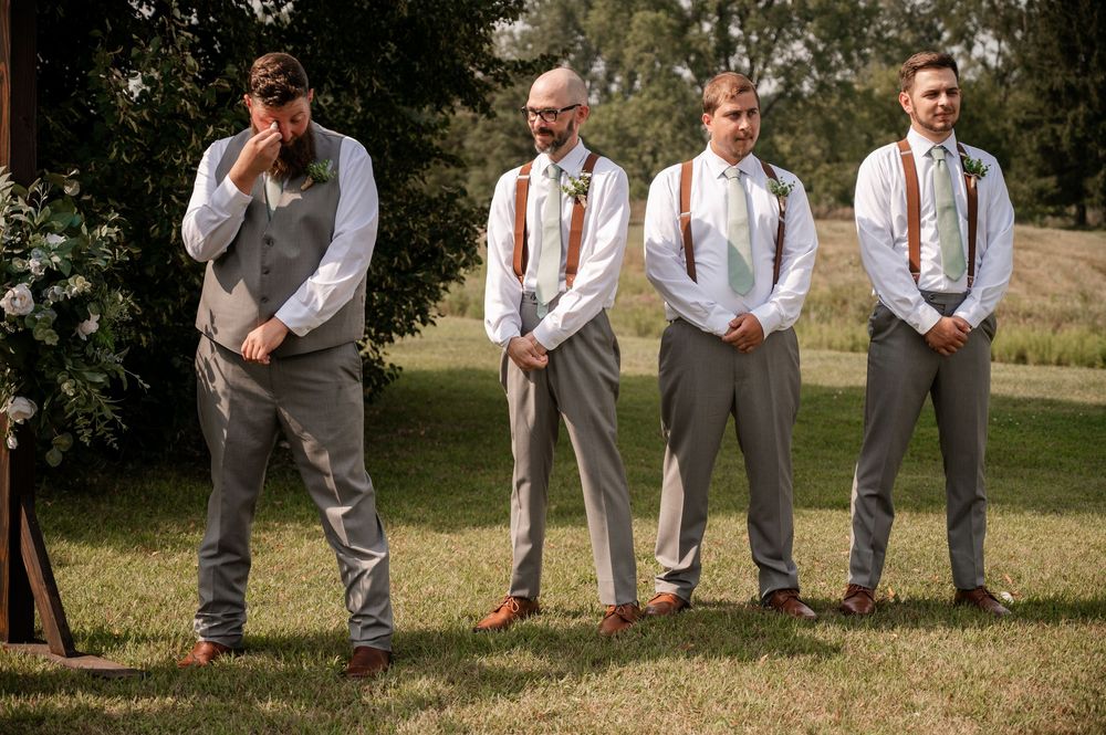 Four groomsmen in gray vests and suspenders standing outdoors at wedding ceremony, one wiping emotional tears.
