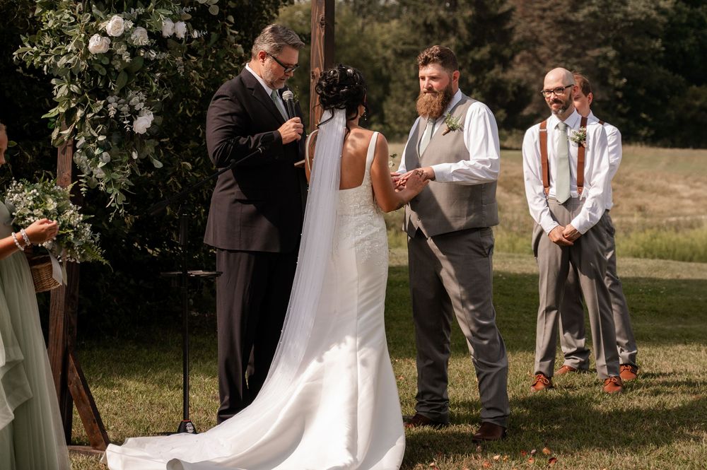 Outdoor wedding ceremony with bride in white gown exchanging vows with groom before officiant and groomsmen.