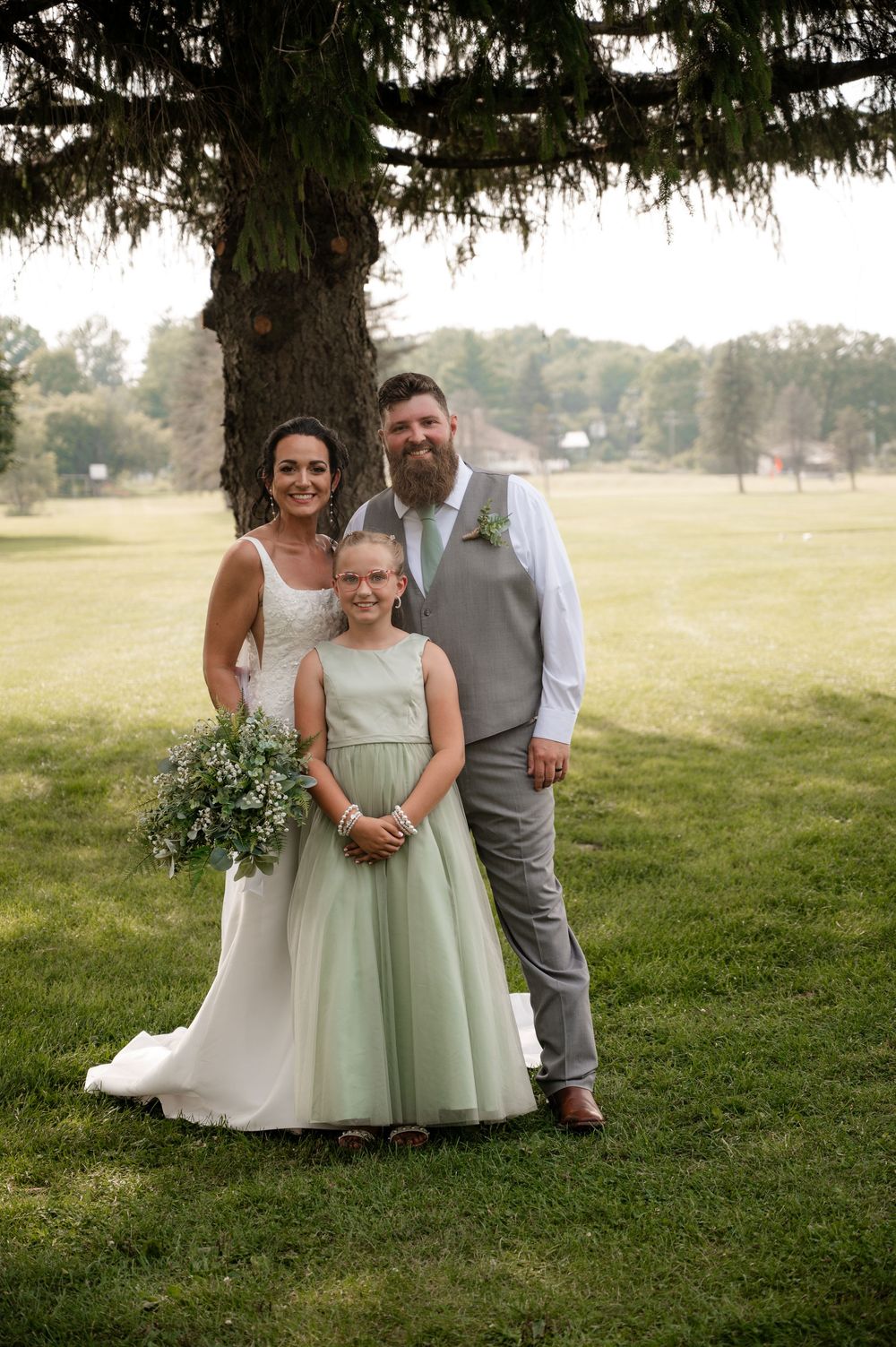 Bride in white gown, groom in gray vest, and young flower girl in sage dress posing under large tree.