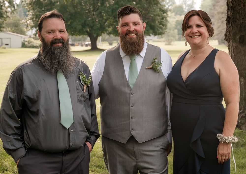 Two bearded men in gray suits with mint ties and a woman in navy dress posing together at outdoor wedding.