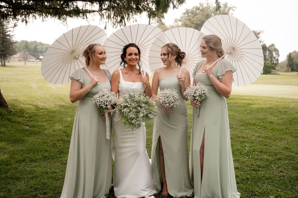 Bride in white gown with three bridesmaids in sage green dresses holding white parasols and floral bouquets outdoors.