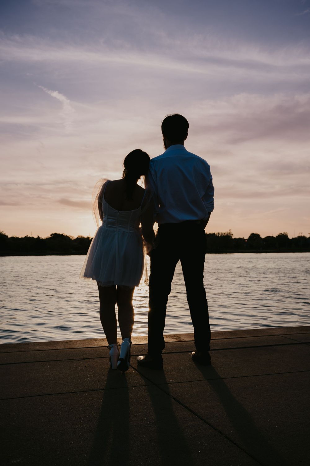 Silhouette of couple standing together on waterfront dock watching a colorful sunset over calm water.