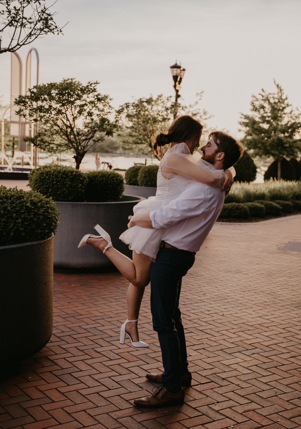 Man lifting woman in white dress for a joyful embrace on a brick patio with greenery at sunset.