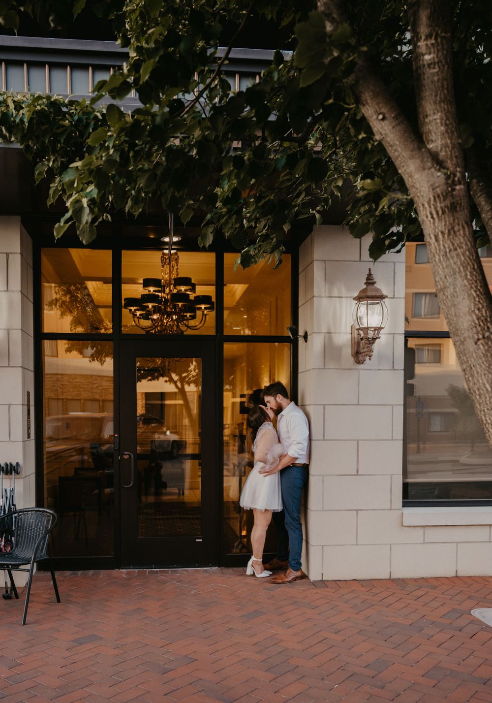 Romantic couple sharing a kiss outside an elegant building entrance with chandelier and brick walkway.