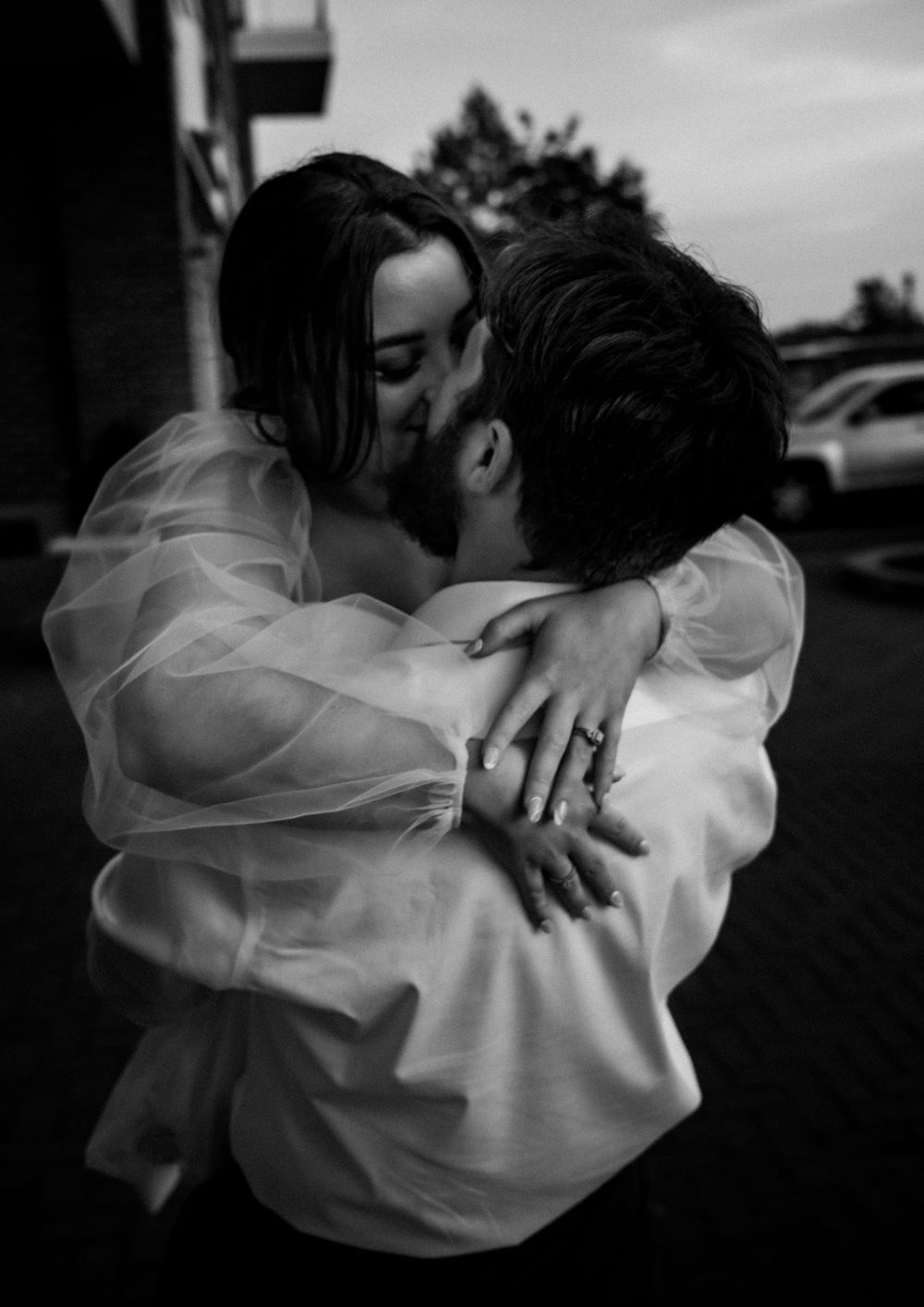 Black and white photo of couple sharing a passionate kiss, woman wearing sheer white gown with ring visible.