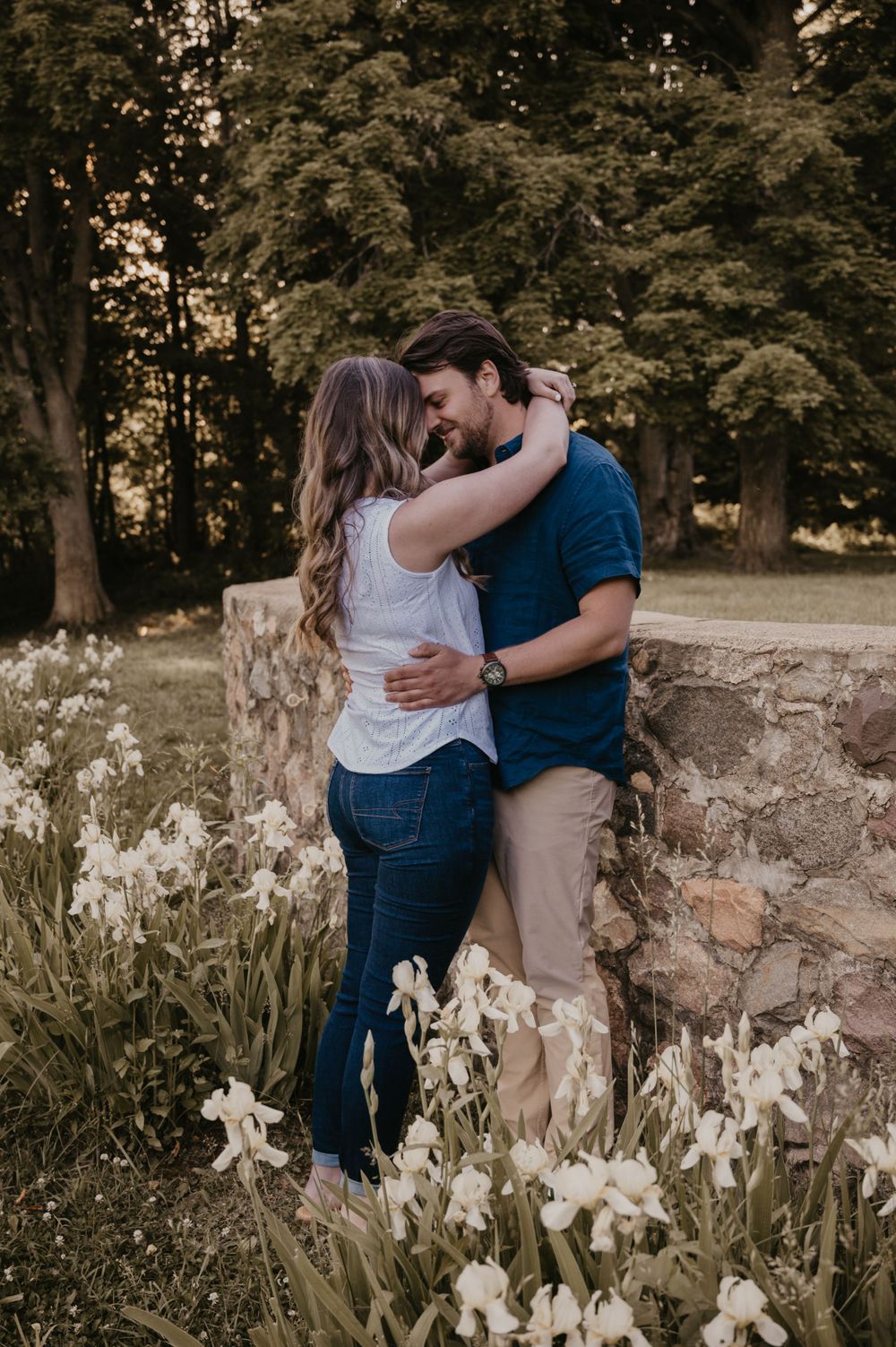 Couple embracing and nearly kissing beside a stone wall surrounded by blooming white iris flowers.