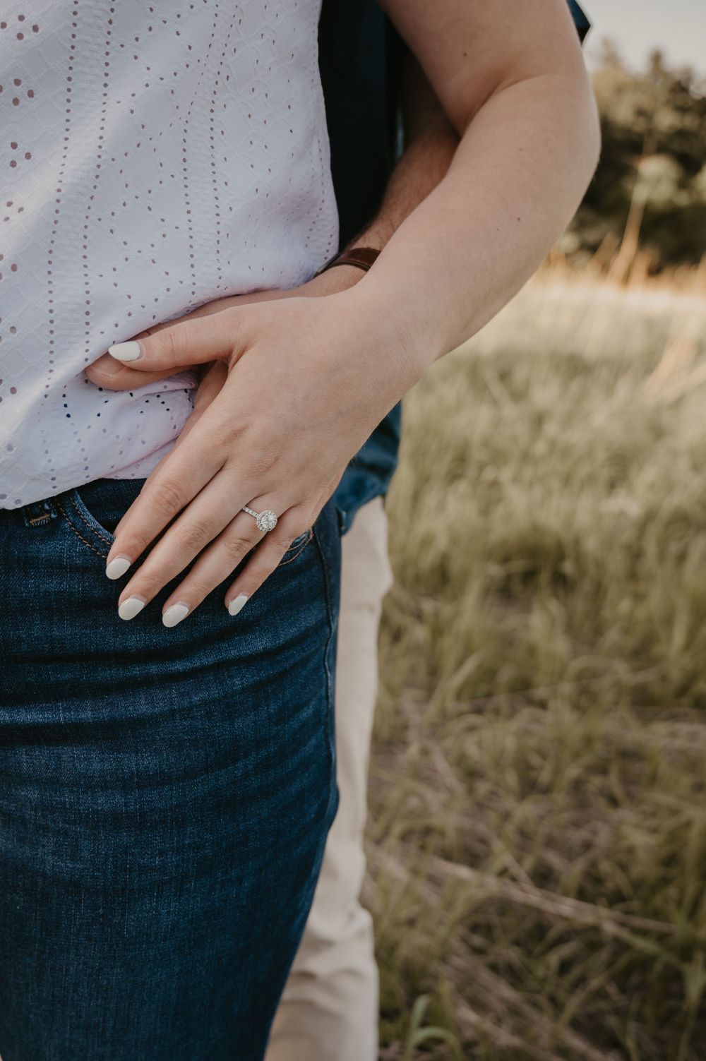 Close-up of woman's hand with elegant engagement ring resting on jeans in a golden field setting.