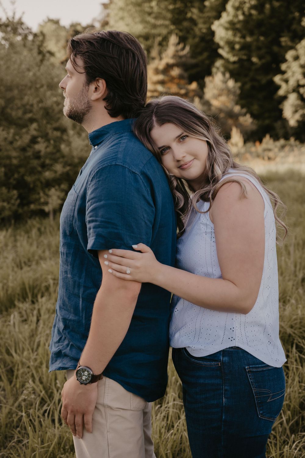 Woman with engagement ring leaning her head on man's back in a lush green meadow at golden hour.