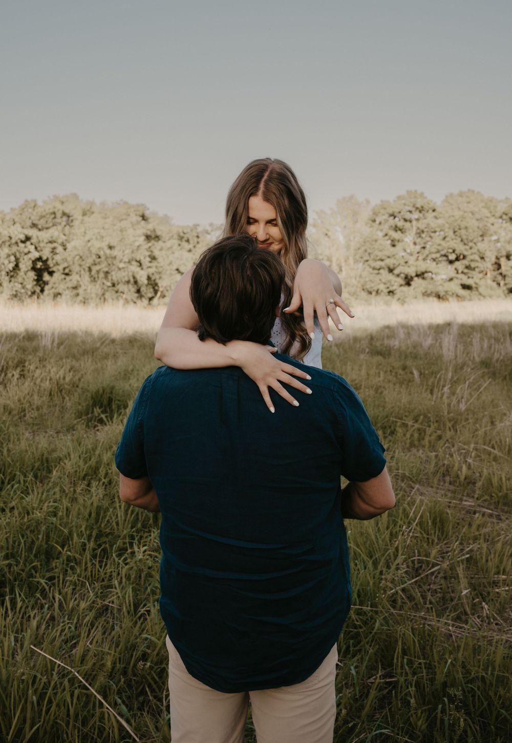 Woman smiling while embracing man from behind in a wide open grassy field during golden hour.