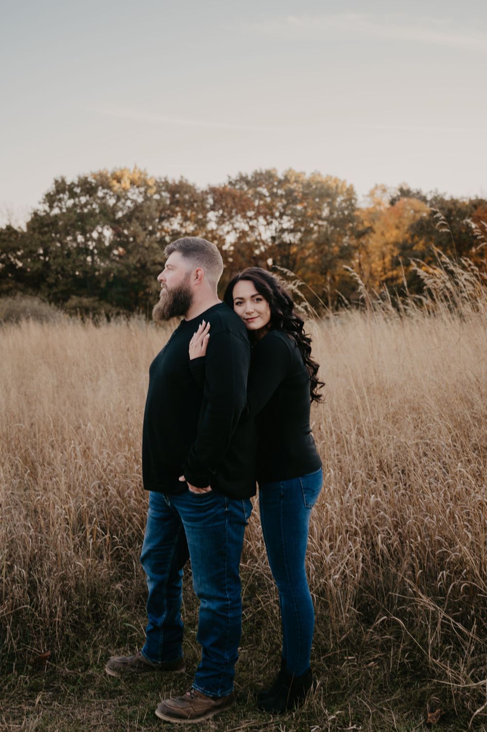 Couple poses romantically in golden autumn field at sunset, woman resting hand on man's back.