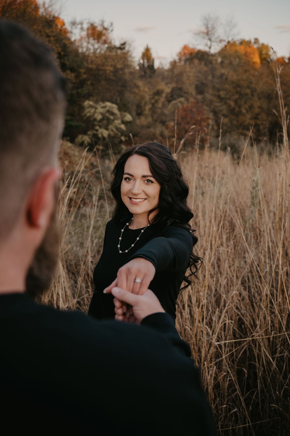Smiling dark-haired woman holds her partner's hand in a golden autumn field.
