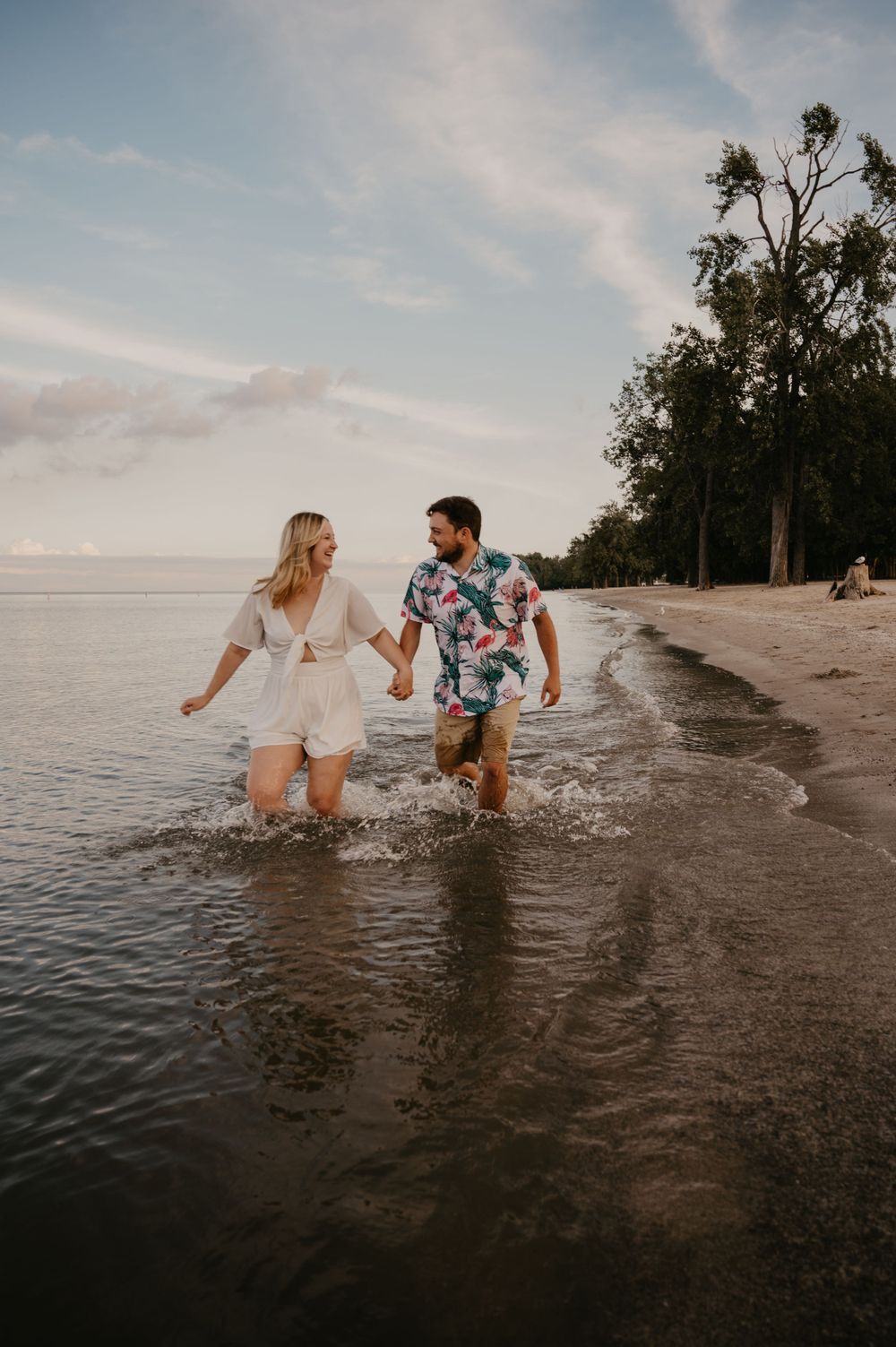 Happy couple holding hands and splashing through shallow water on a sandy beach at golden hour.