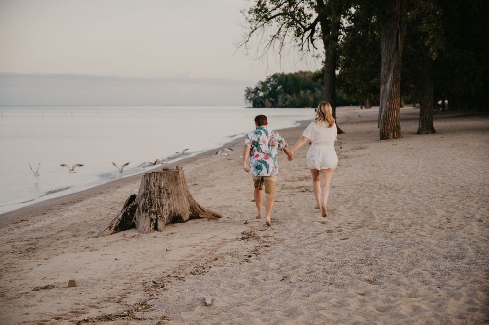 Couple holding hands running along sandy beach shoreline at sunset near tall trees.