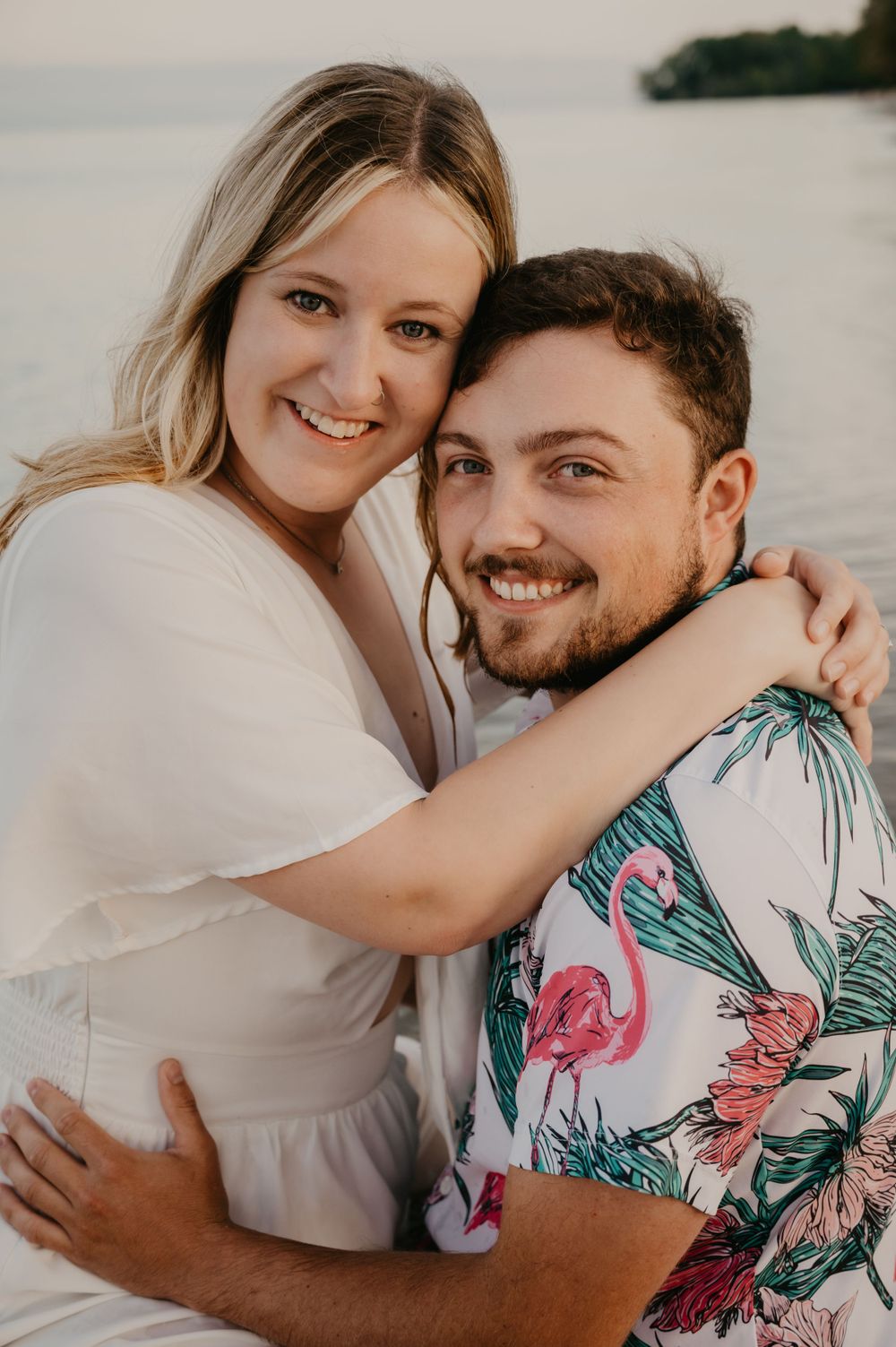 Smiling couple embracing near water, woman in white dress hugging man in tropical flamingo shirt at sunset.