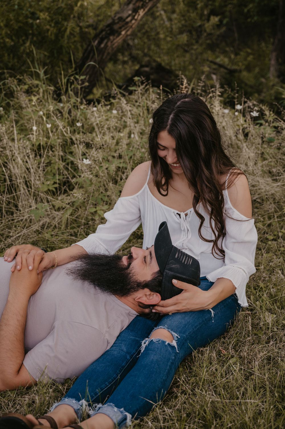 Woman in white top smiling down at bearded man lying in her lap in a lush green meadow.