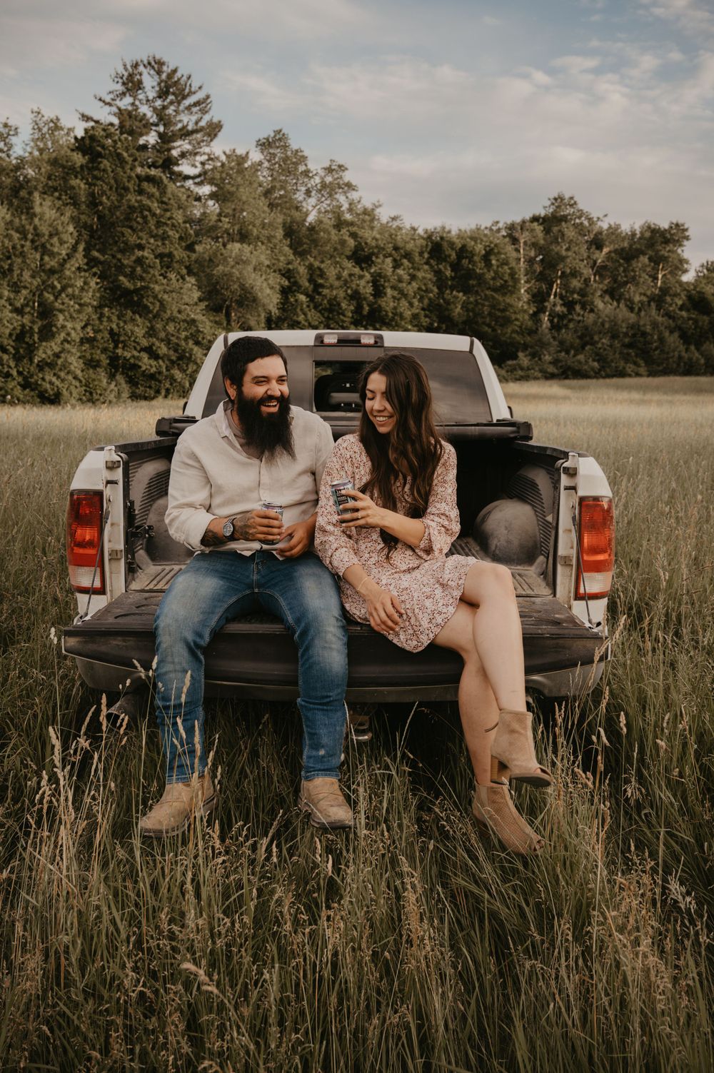 Smiling couple sitting on truck tailgate in tall grass field, holding beverages during outdoor photo session.