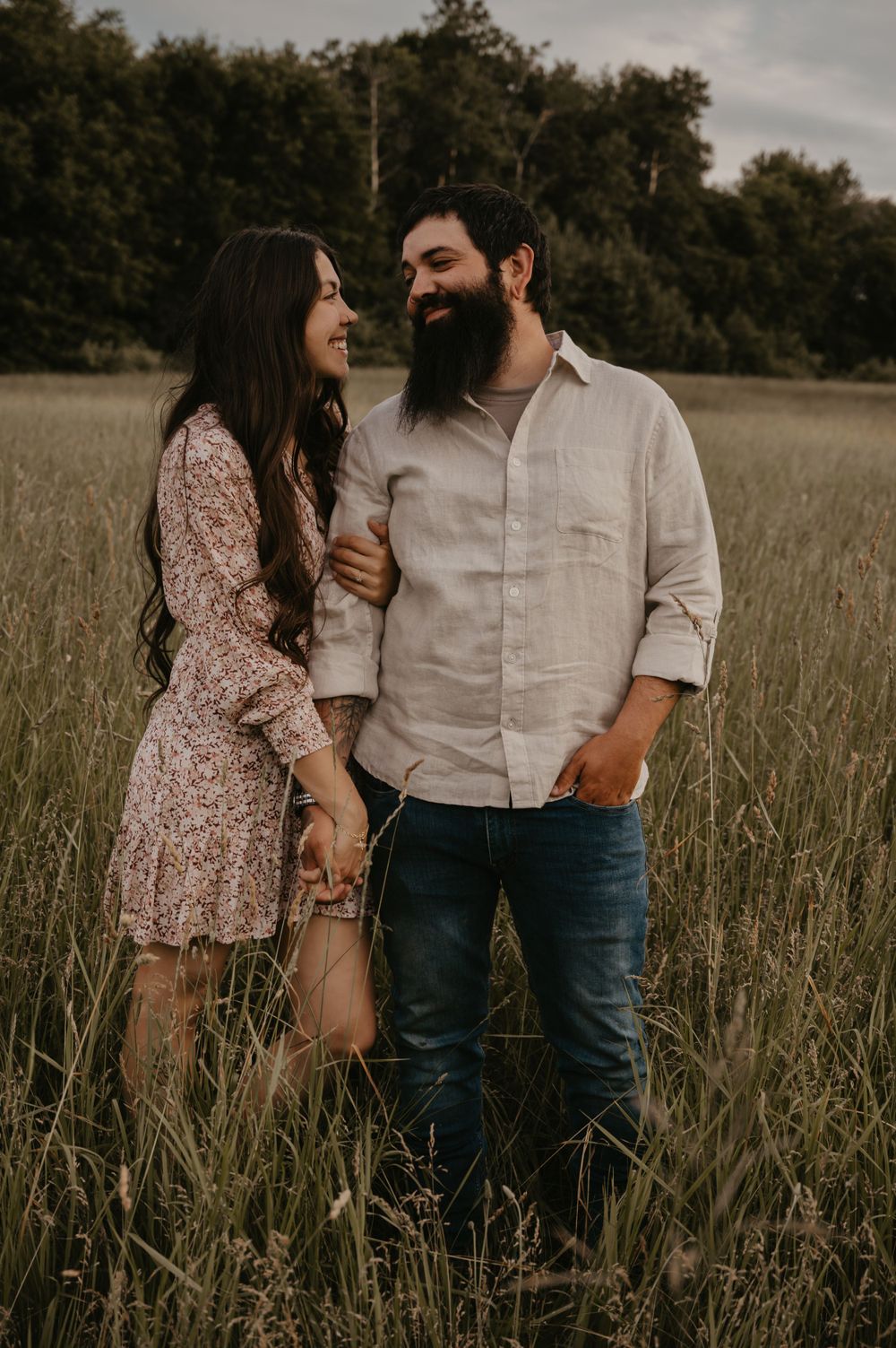 Laughing couple holding hands while standing in tall golden grass field, woman in floral dress.