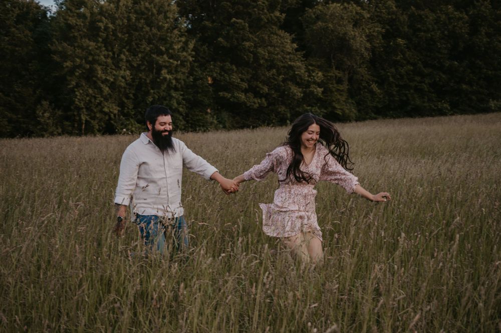 Joyful couple running hand in hand through tall grass field surrounded by trees at dusk.