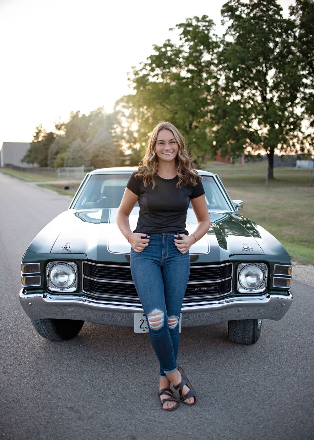 Confident young woman in jeans leans against a classic muscle car at golden hour.