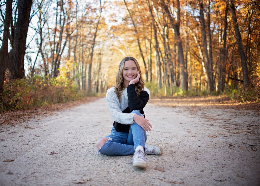 Smiling teenage girl sits on a dirt path surrounded by colorful autumn foliage.