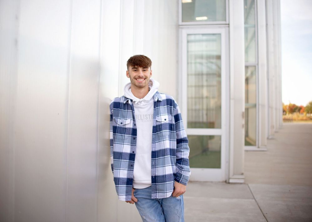 Smiling young man in a plaid flannel shirt leans against a modern building exterior.