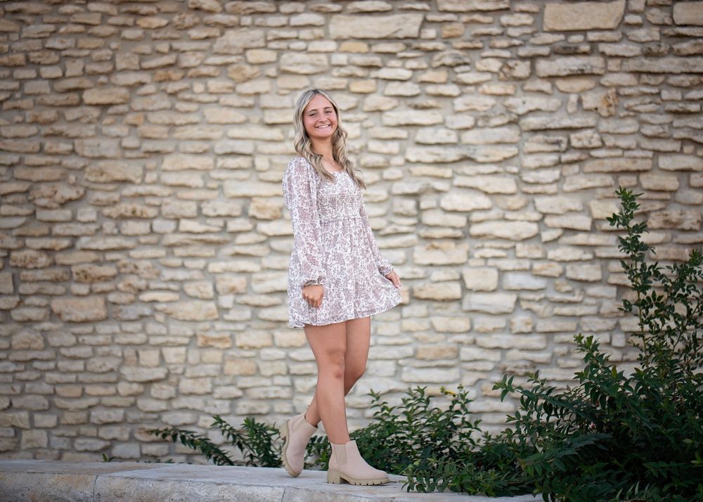 Young woman in a floral mini dress and tan boots poses against a rustic stone wall.