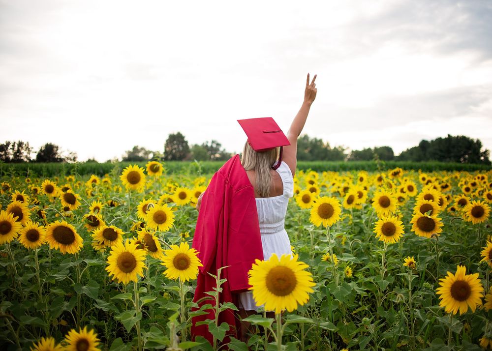 Graduate in red cap and gown raises peace sign in a vibrant sunflower field.
